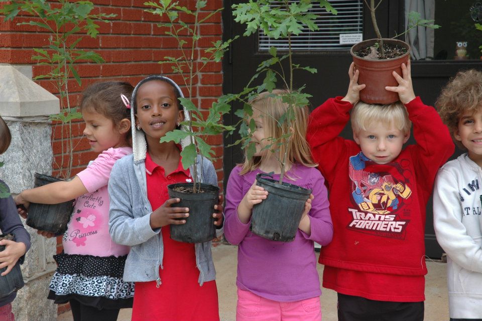 Group of elementary aged students holding tree seedlings in pots in front of their school.