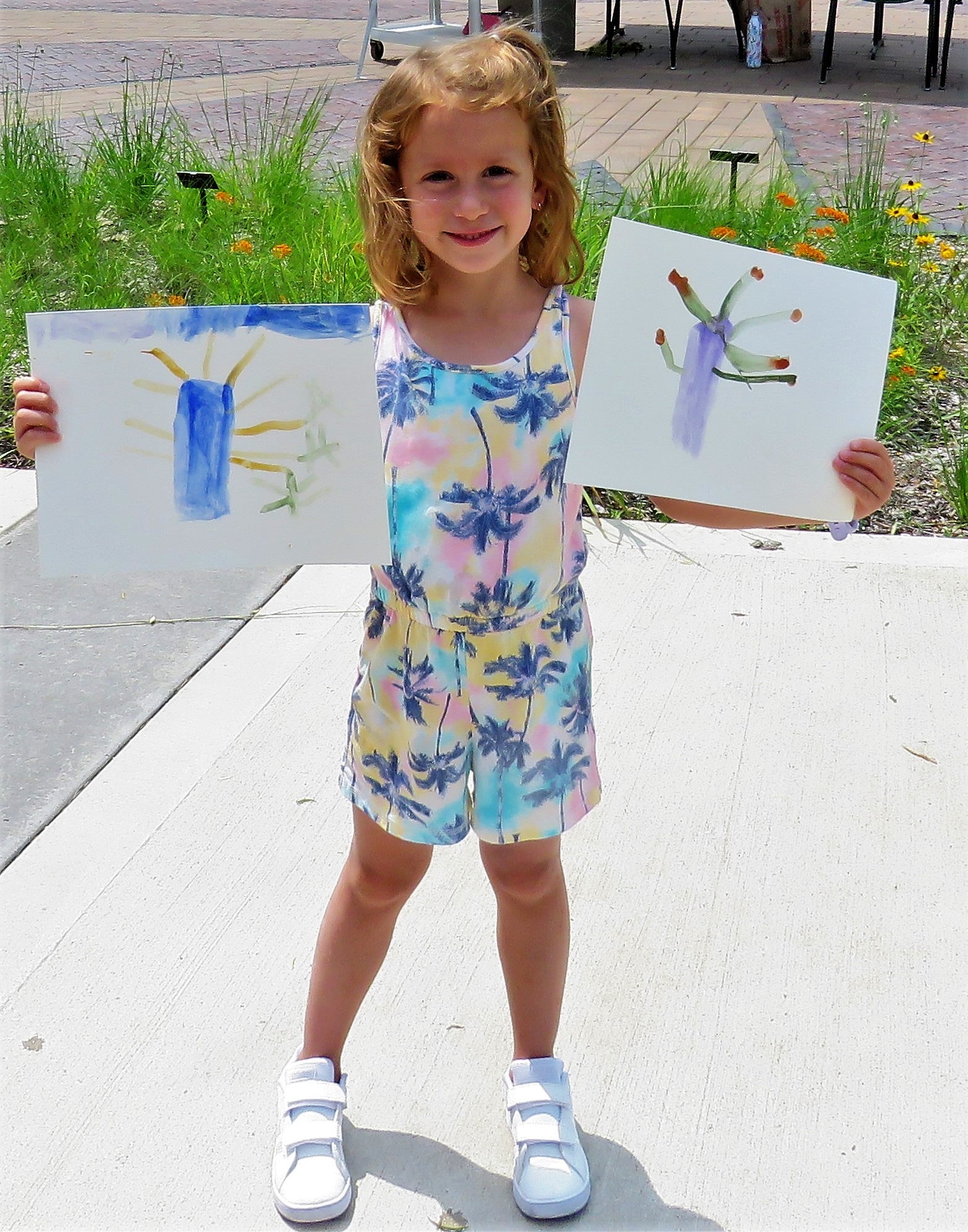 Young girl with strawberry blonde hair holding two botanical art projects she created featuring trees. She is standing in a garden.